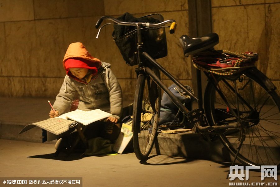 鄭州中原路京廣鐵路大橋下涵洞的馬路牙子邊，一位戴著紅色辣椒帽的小男孩趴在一個小馬扎上，上面鋪著一張車墊，在微弱的燈光下寫作業(yè)