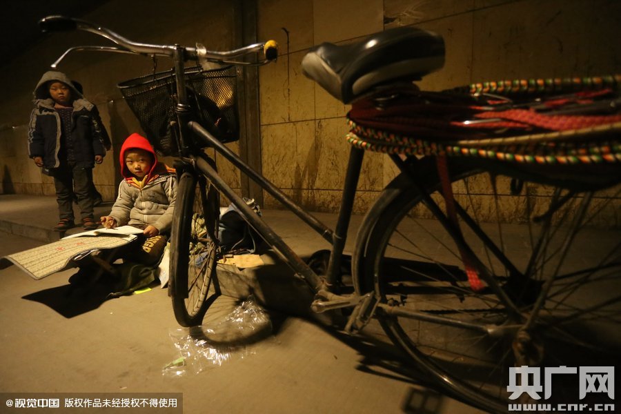 2015年11月8日晚，鄭州，7歲男孩涵洞橋下陪父母練攤，路邊燈下坐小馬扎上寫作業(yè)。